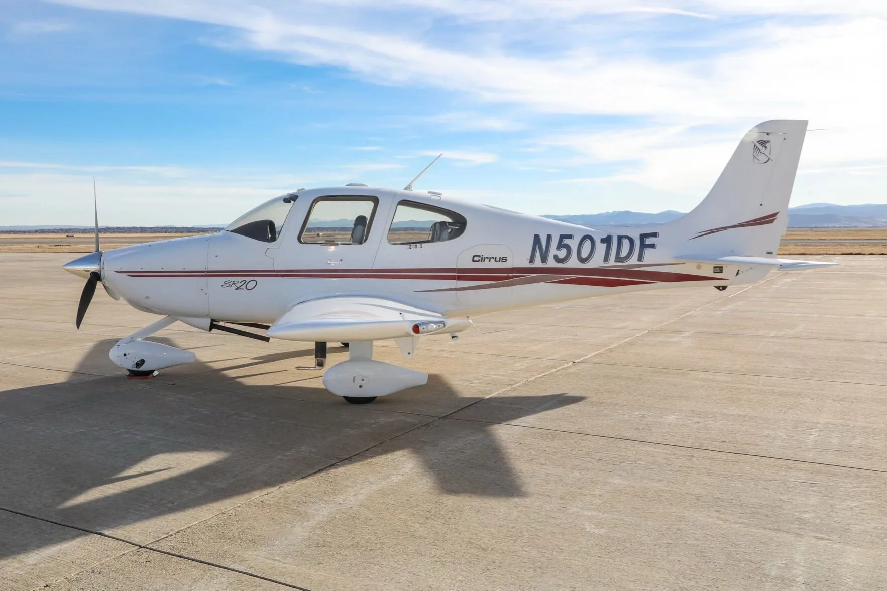 A white Cirrus SR20 aircraft with red stripes, tail number N501DF, parked on an airfield under a blue sky.
