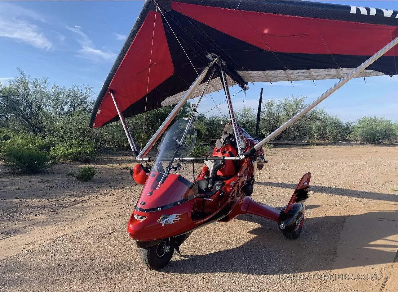 A red weight-shift control aircraft with a black and red wing is parked on a dirt path outdoors.