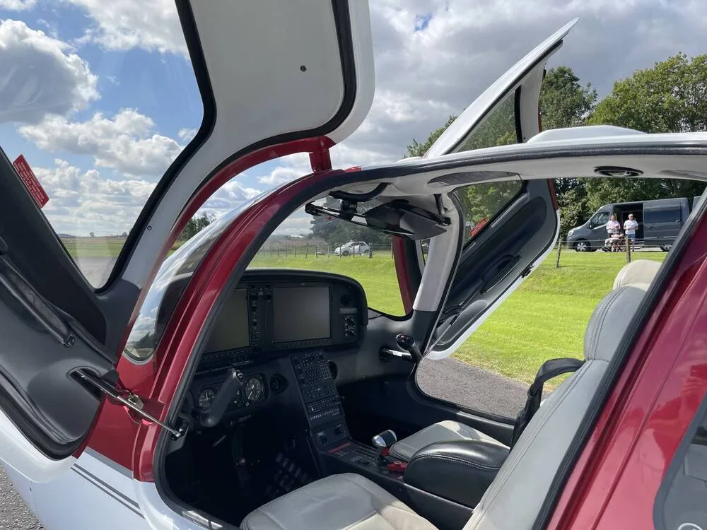 The interior of a Cirrus SR22 aircraft shows the light-colored seats and part of the cockpit with the doors open.