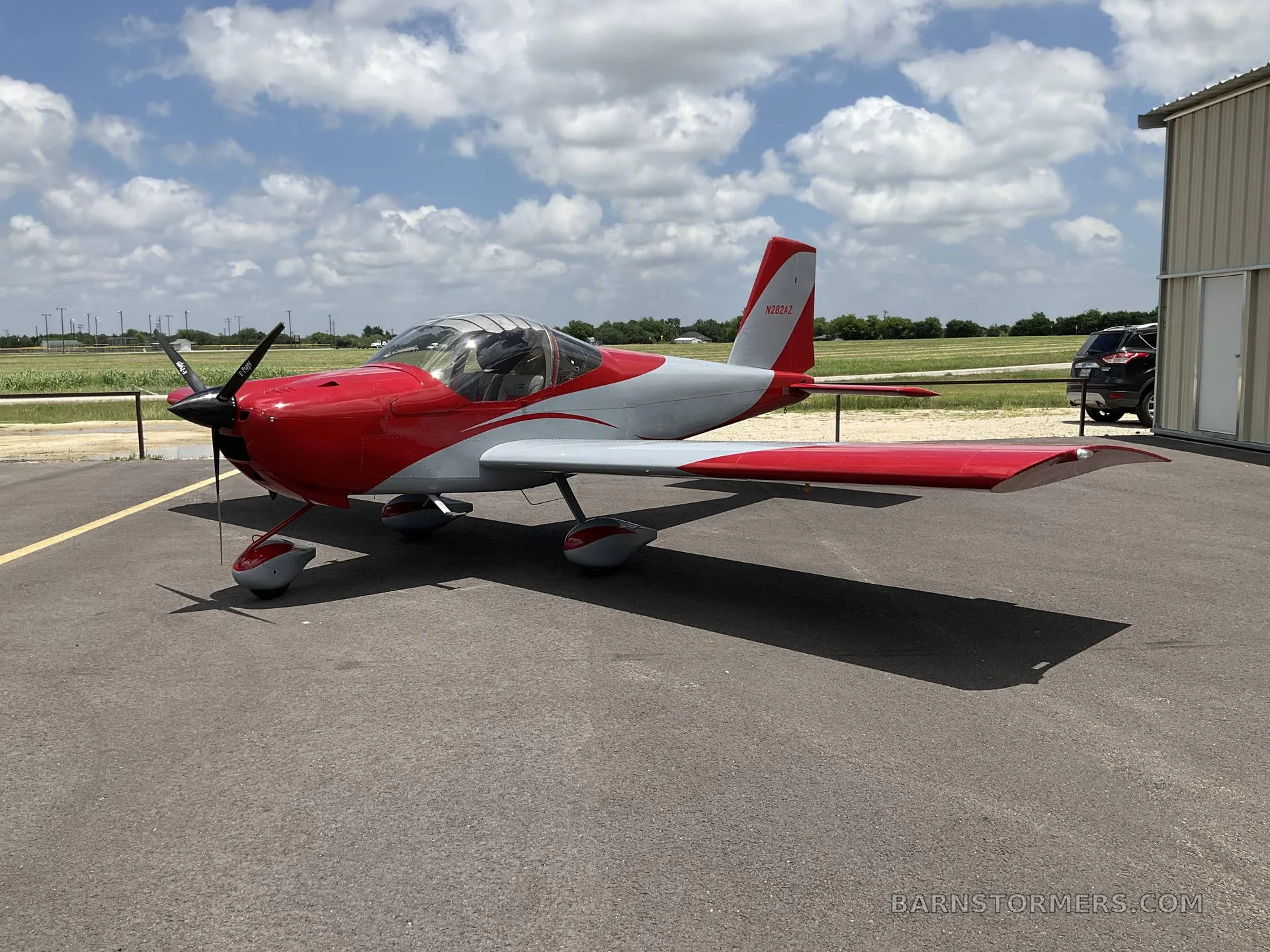 A red and grey single-engine aircraft with a propeller, parked on an asphalt surface under a partly cloudy sky.