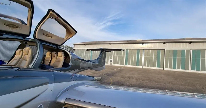 A dark blue and silver Diamond DA62 aircraft with its gull-wing doors open, revealing light-colored seats, parked in front of hangars.