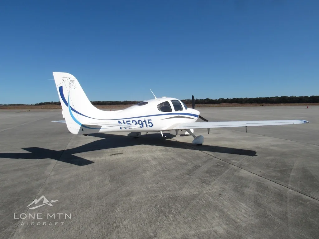 A white Cirrus SR20 aircraft with blue and grey stripes is seen from the rear-left on a concrete ramp.