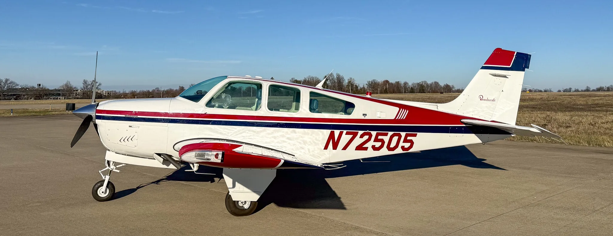 Side-right view of a white Beechcraft Bonanza with red and blue stripes, showing registration N72505, on an airfield.