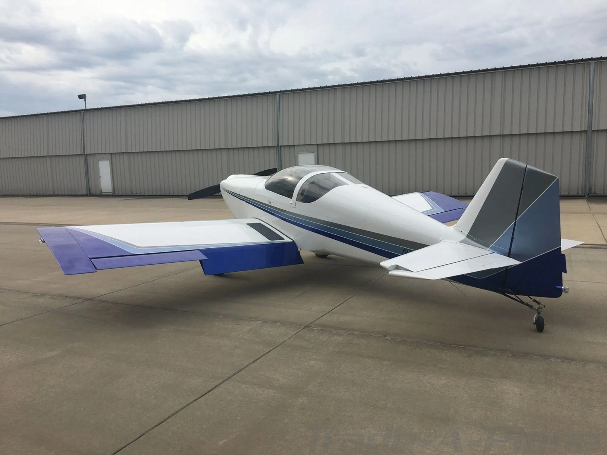 A white Van's RV-6 aircraft with blue and gray stripes parked on an airfield in front of a hangar under a cloudy sky.