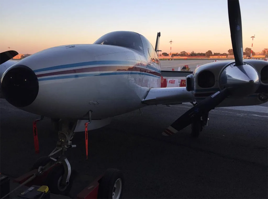 Front view of a white twin-engine aircraft with blue and red stripes, parked on an airfield with a tow bar attached, at sunset.