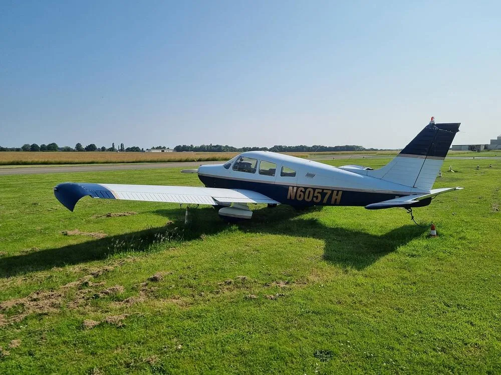 A Piper PA-28 Cherokee aircraft, registration N6057H, with blue and white livery, parked on grass beside a runway on a sunny day.