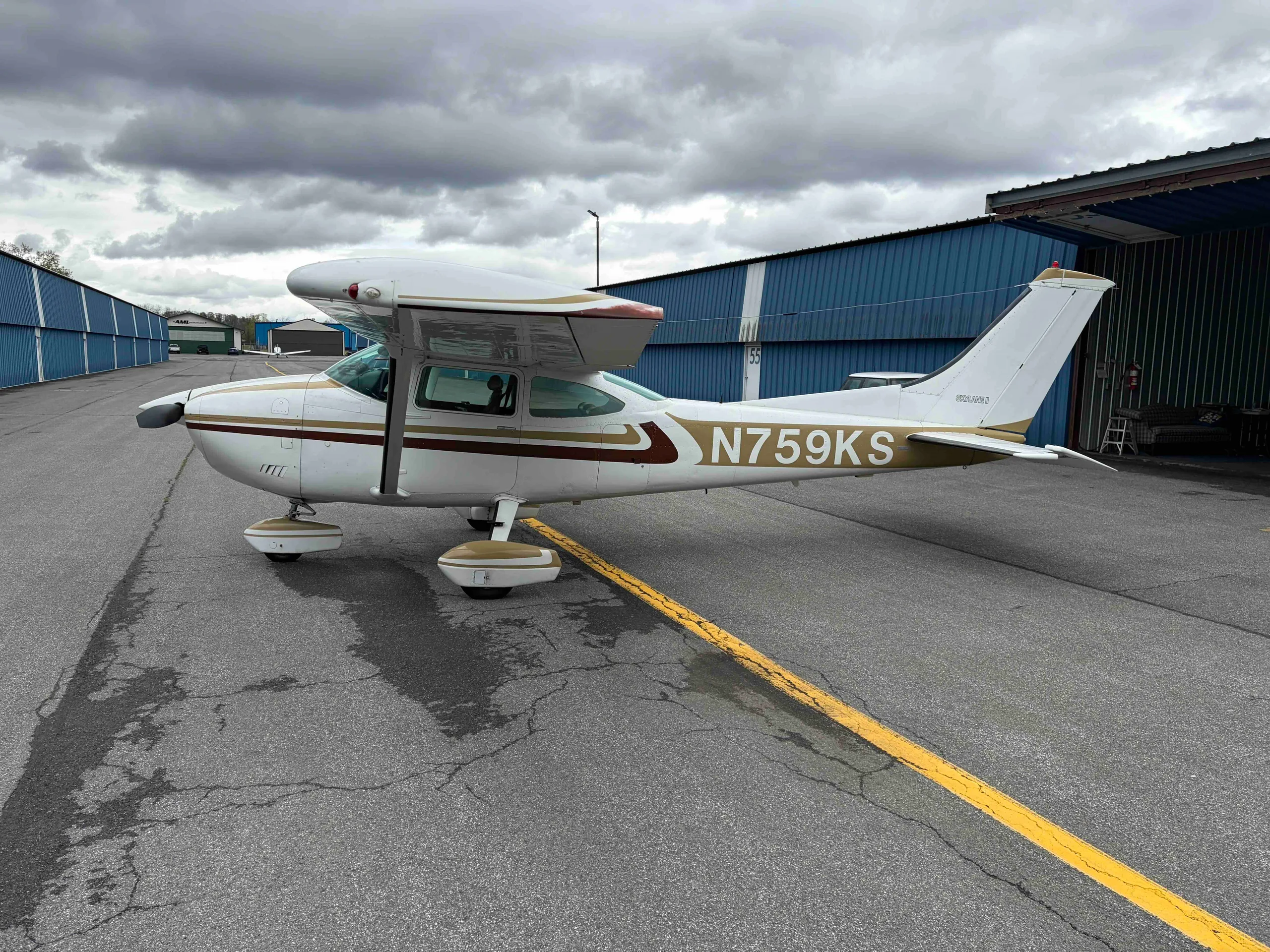 A side view of the white Cessna 182 Skylane N759KS with gold and brown stripes, parked on an airport tarmac.