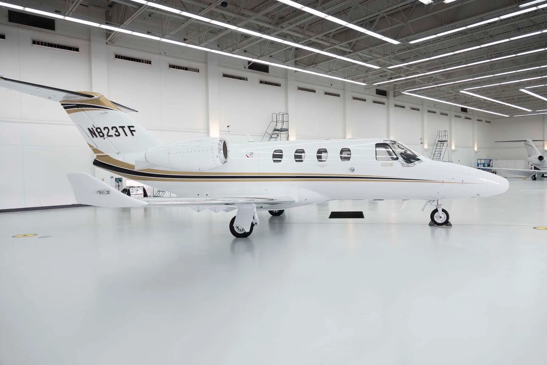 A side view of a white Cessna Citation M2 jet with gold and black stripes, parked in a hangar.