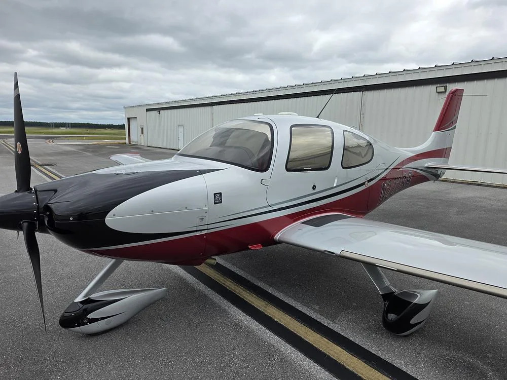A white, red, and black single-engine aircraft with a propeller on a tarmac, viewed from the front-left.