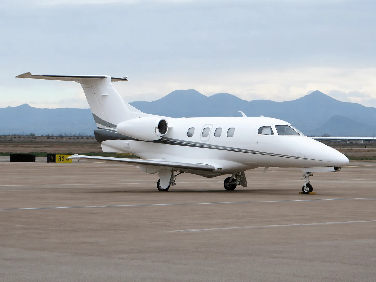 A white business jet with grey stripes is parked on an airport tarmac with mountains in the background under a cloudy sky.