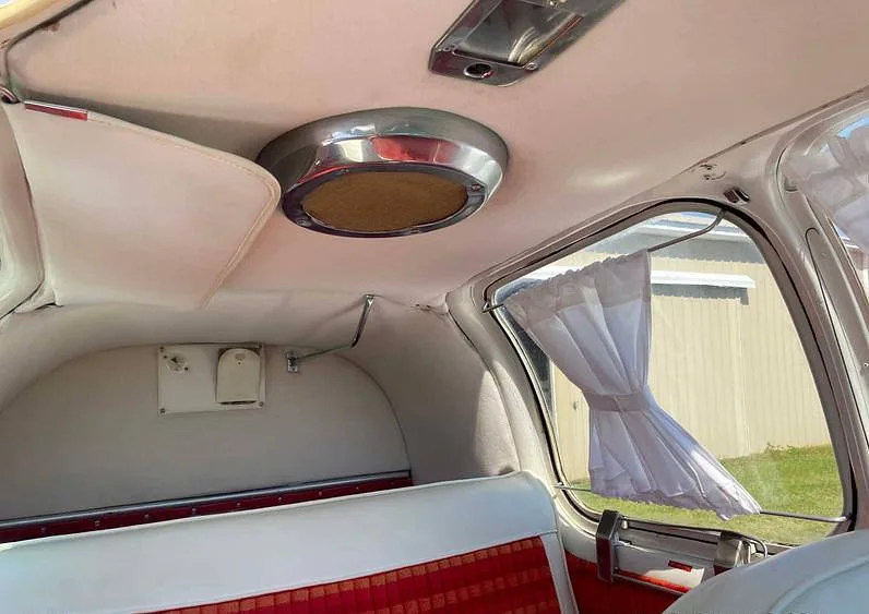 The aircraft's interior cabin shows white seats with red patterned inserts, a window with a white curtain, and ceiling details.
