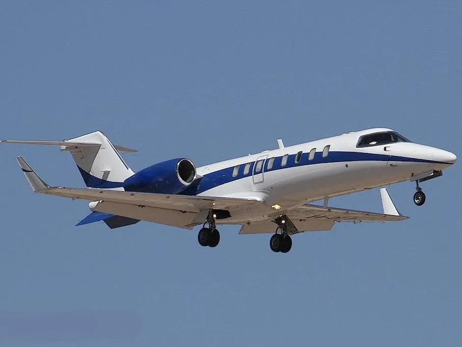 A white and blue business jet with landing gear extended, seen from below against a clear blue sky.