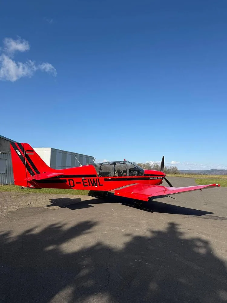 A red GR 400-200 light aircraft with black stripes parked on an airfield tarmac next to a hangar.