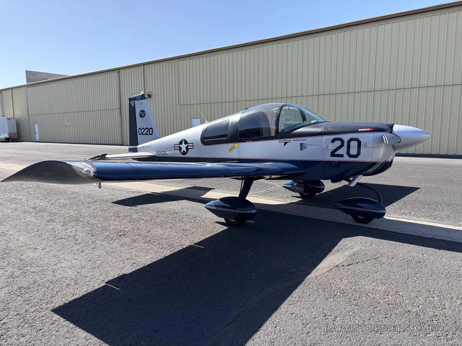 A silver and blue Grumman American AA-5 Traveler aircraft with a military-style paint scheme, parked on an asphalt ramp next to a hangar.