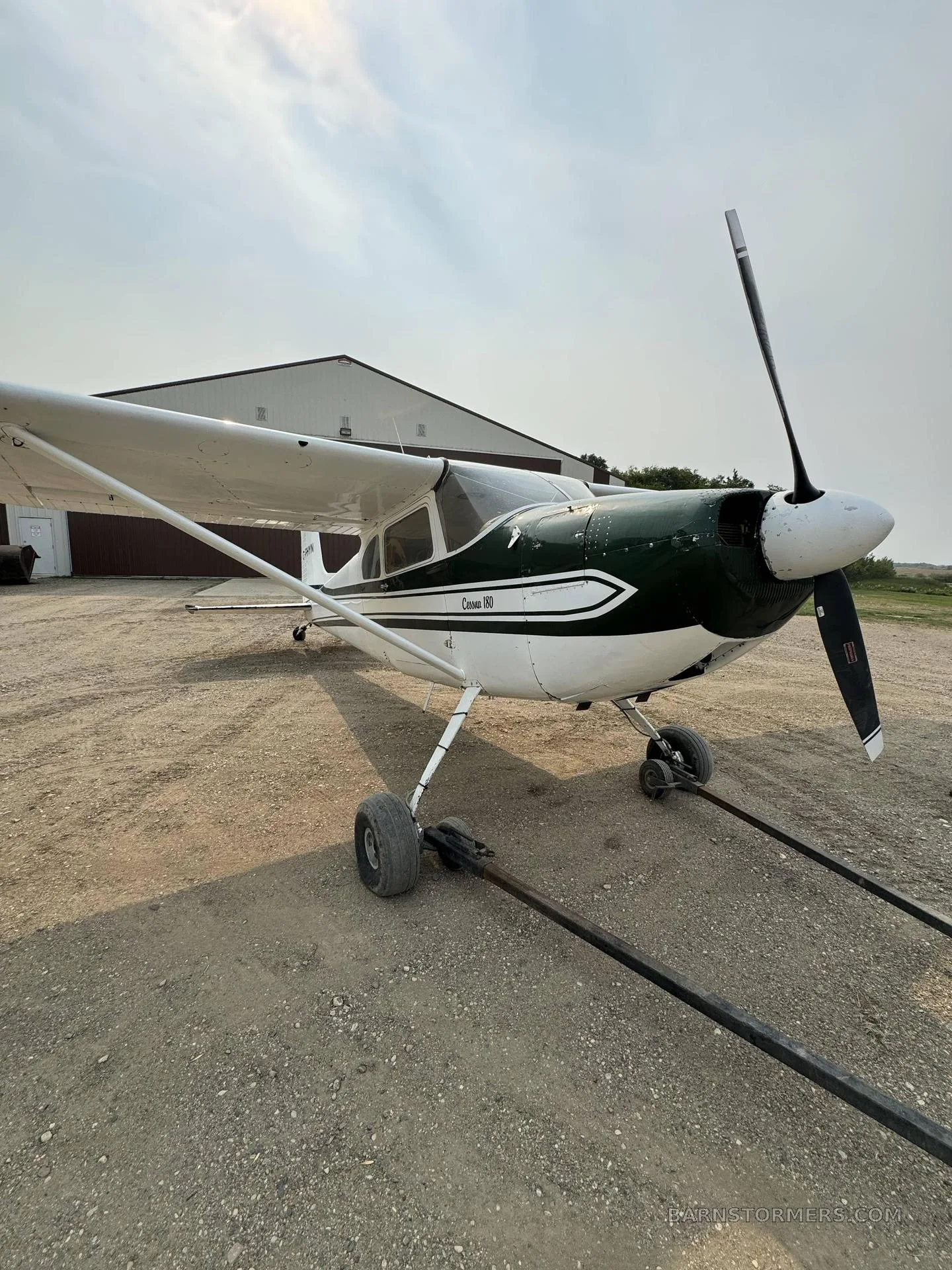 A front-left view of a white and green Cessna 180 aircraft parked on a gravel lot in front of a hangar.