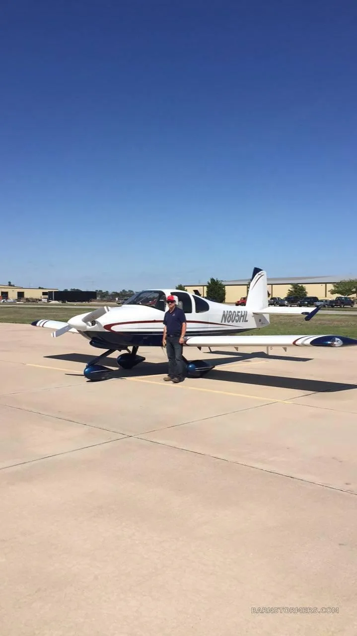 A man stands next to a white and blue single-engine aircraft, N805HL, on an airfield under a clear blue sky.