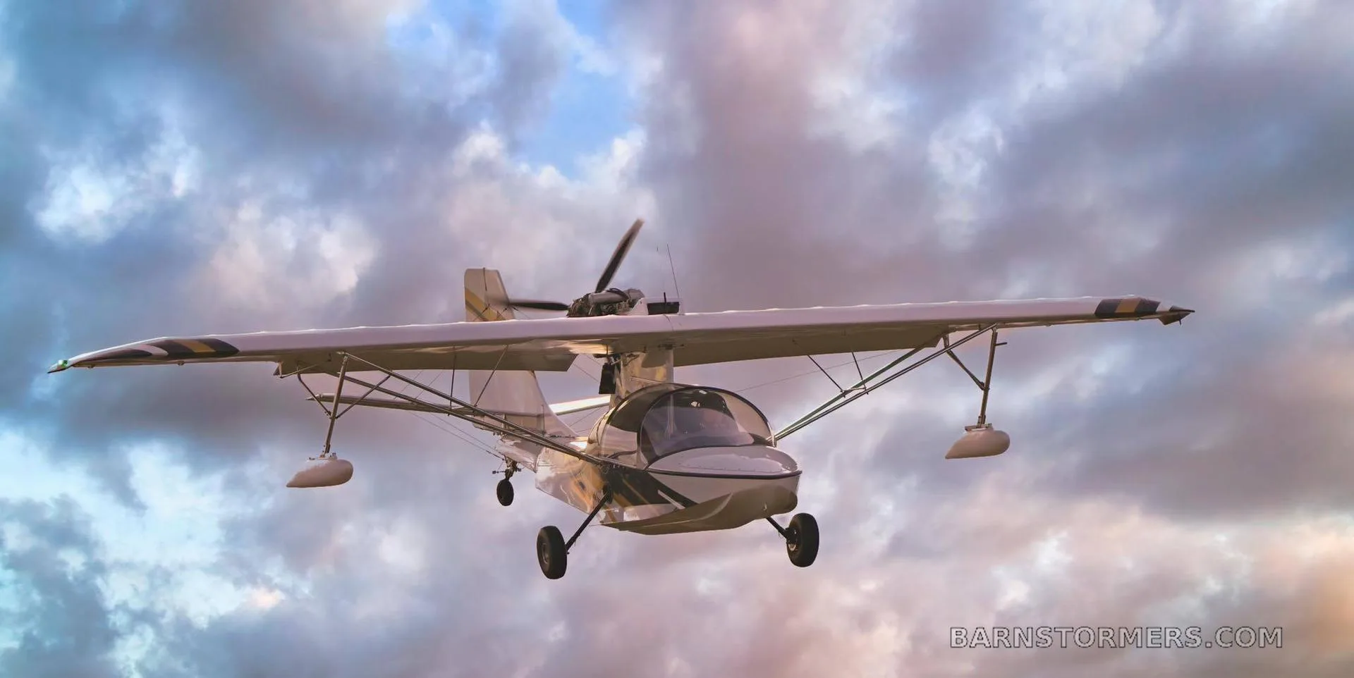 A white and gold amphibious light sport aircraft with floats and wheels in flight against a cloudy sky.
