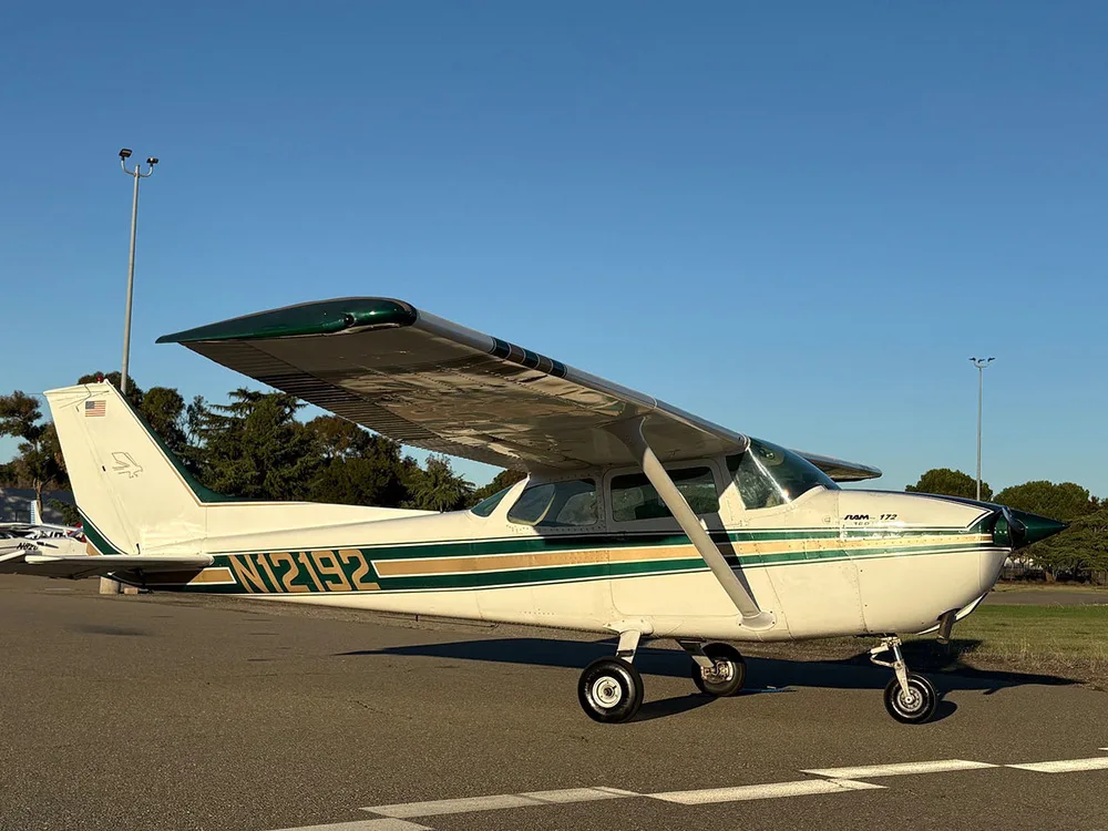 A white Cessna 172 with green and gold stripes parked on an airfield with trees in the background.