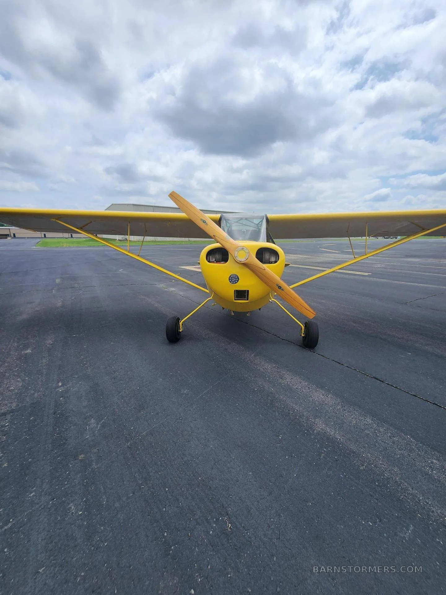 A head-on view of a yellow single-engine aircraft with a wooden propeller, parked on an asphalt tarmac.