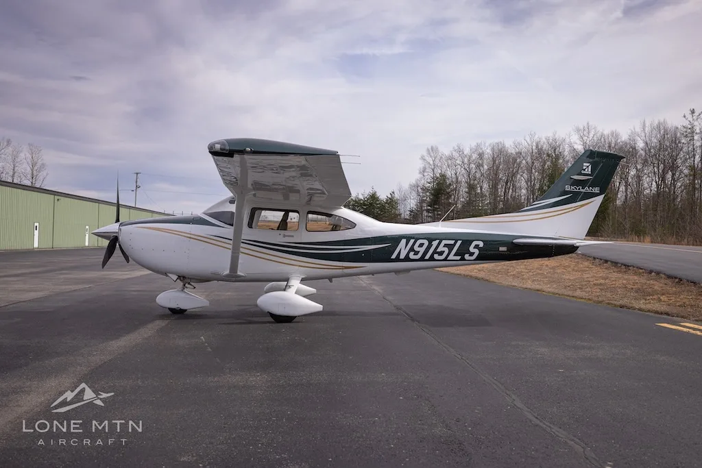 A side view of a white and dark green Cessna Skylane N915LS with gold stripes and flaps extended, parked on an asphalt ramp.