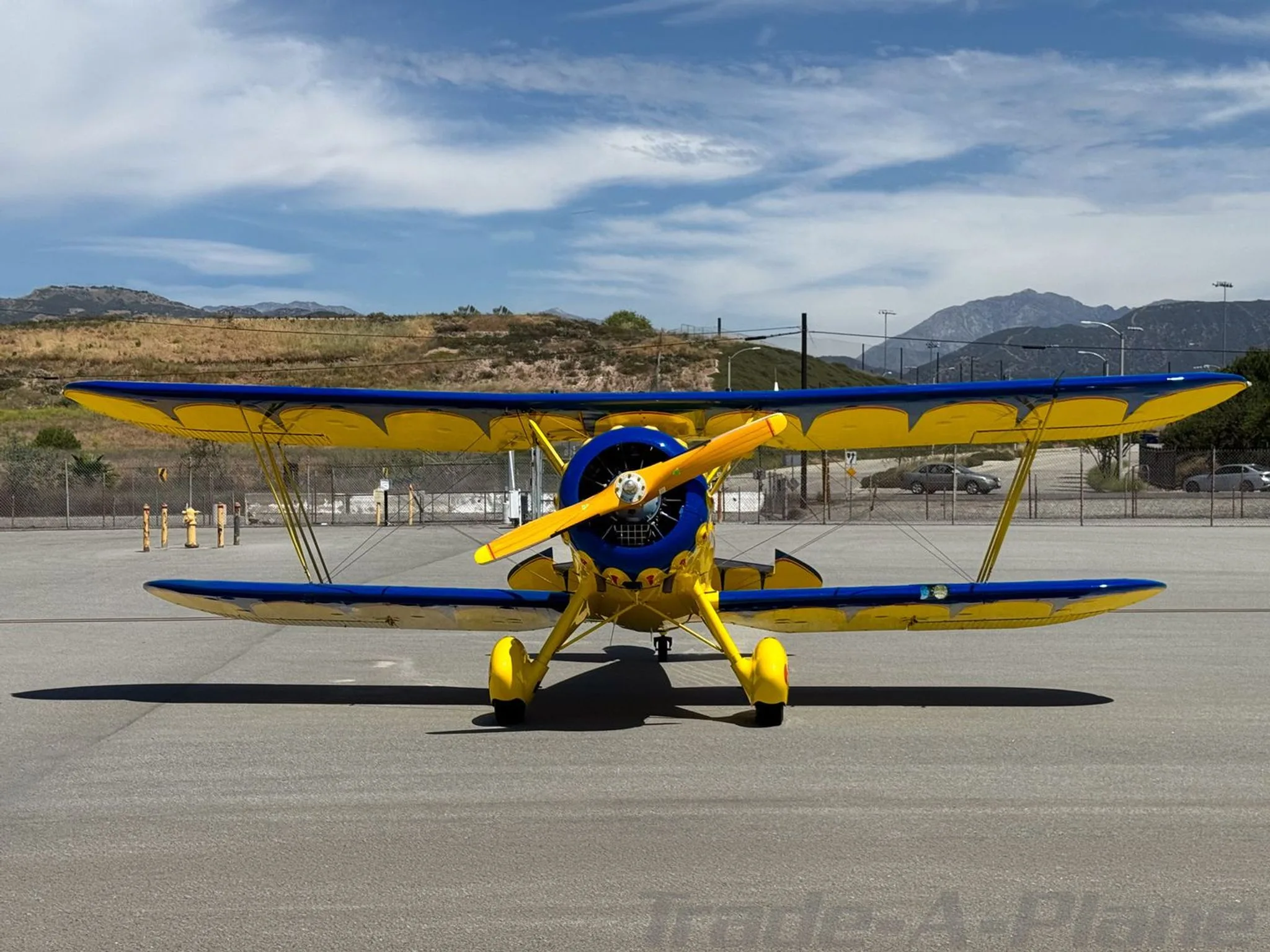 A head-on view of a yellow and blue biplane with a wooden propeller, parked on an airfield.
