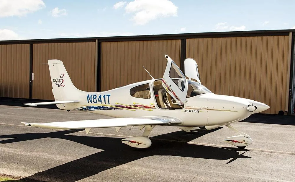 A white Cirrus SR20 aircraft with open doors and blue and red accent stripes parked next to a hangar.