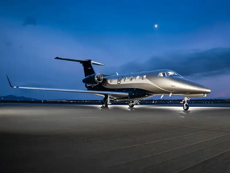 A sleek silver and dark blue private jet is parked on an airfield at dusk, with city lights visible in the background.