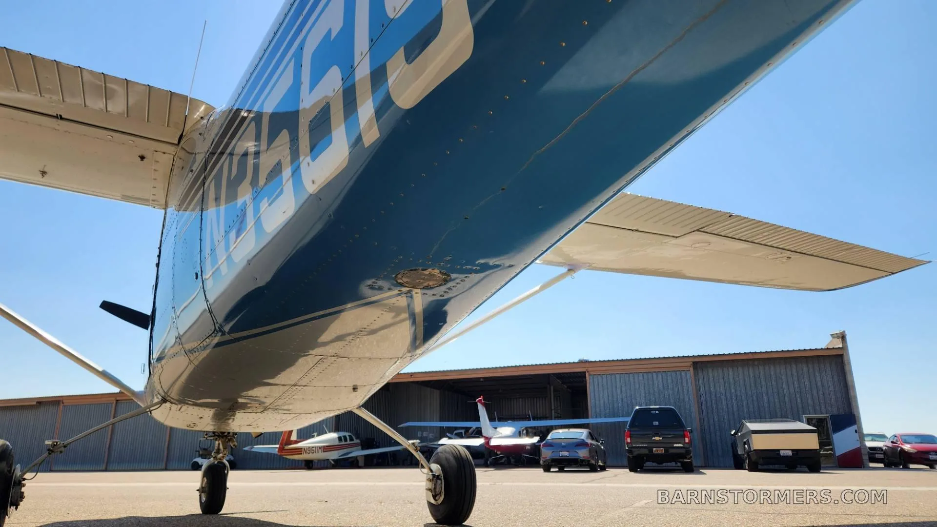 A low-angle view of the underside of a blue and white aircraft's wing and fuselage, parked on an airfield.
