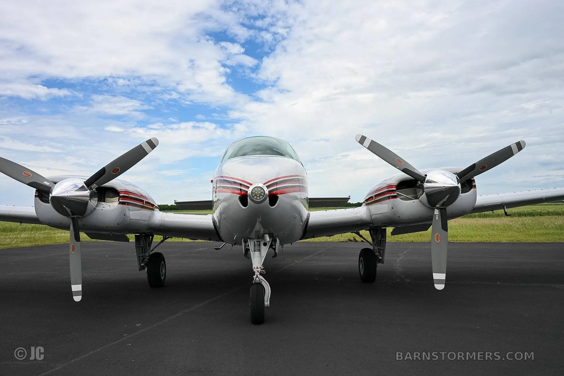 A head-on view of a white and red twin-engine propeller aircraft parked on an asphalt runway under a cloudy sky.
