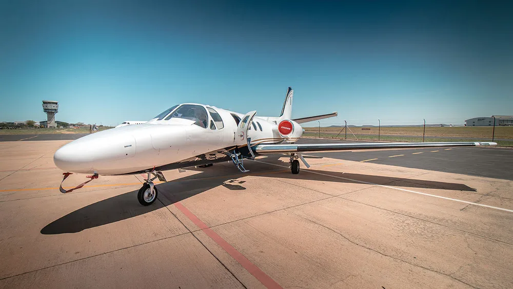 A white Cessna Citation jet with gold and blue stripes parked on an airfield under a clear blue sky.