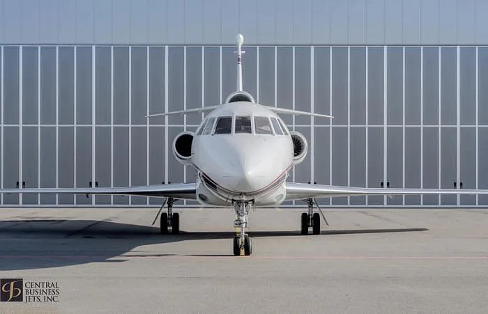 A white business jet with red and black stripes on its nose, seen from the front, parked on an tarmac in front of a modern hangar.