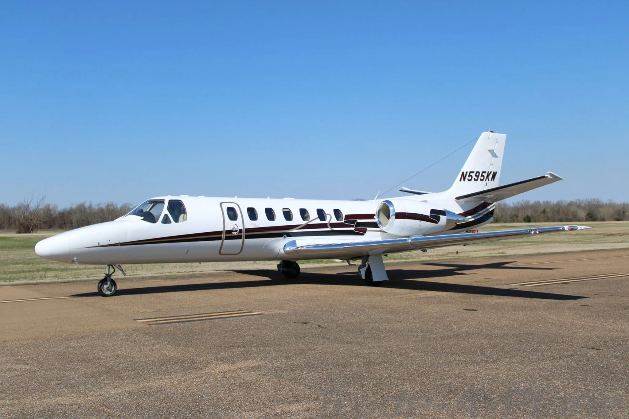 A white Cessna Citation Encore business jet with dark brown and gold stripes on the tarmac under a clear blue sky.