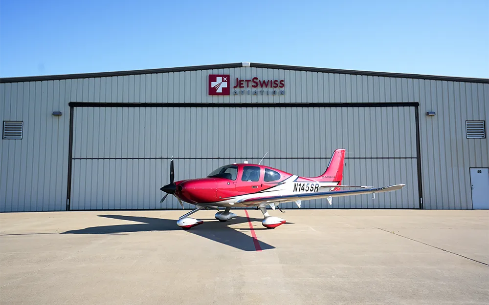 A red and white Cirrus SR22 aircraft with registration N145SR parked on an tarmac in front of a JetSwiss Aviation hangar.