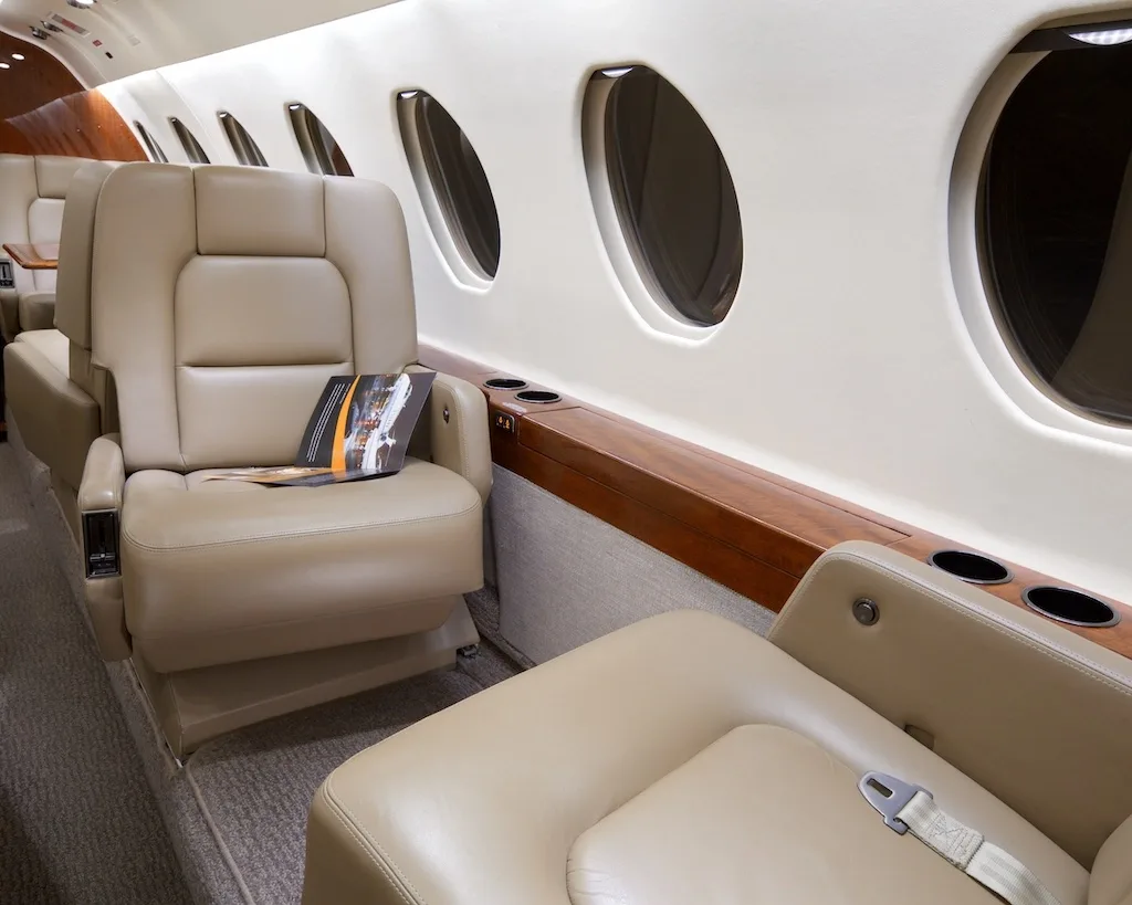 Two beige leather seats in a private jet cabin, with a magazine on one seat and a wooden side panel with cup holders.