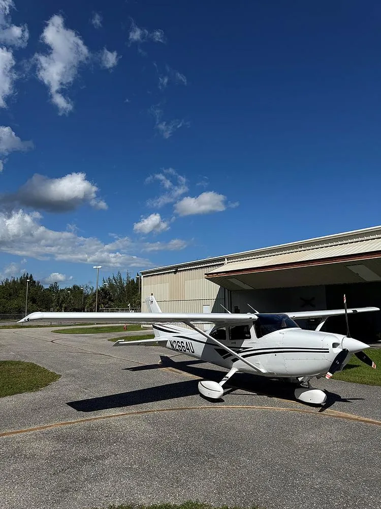 Wide shot of a white Cessna 182 Skylane with black and brown stripes, parked on an asphalt ramp with hangars in the background.