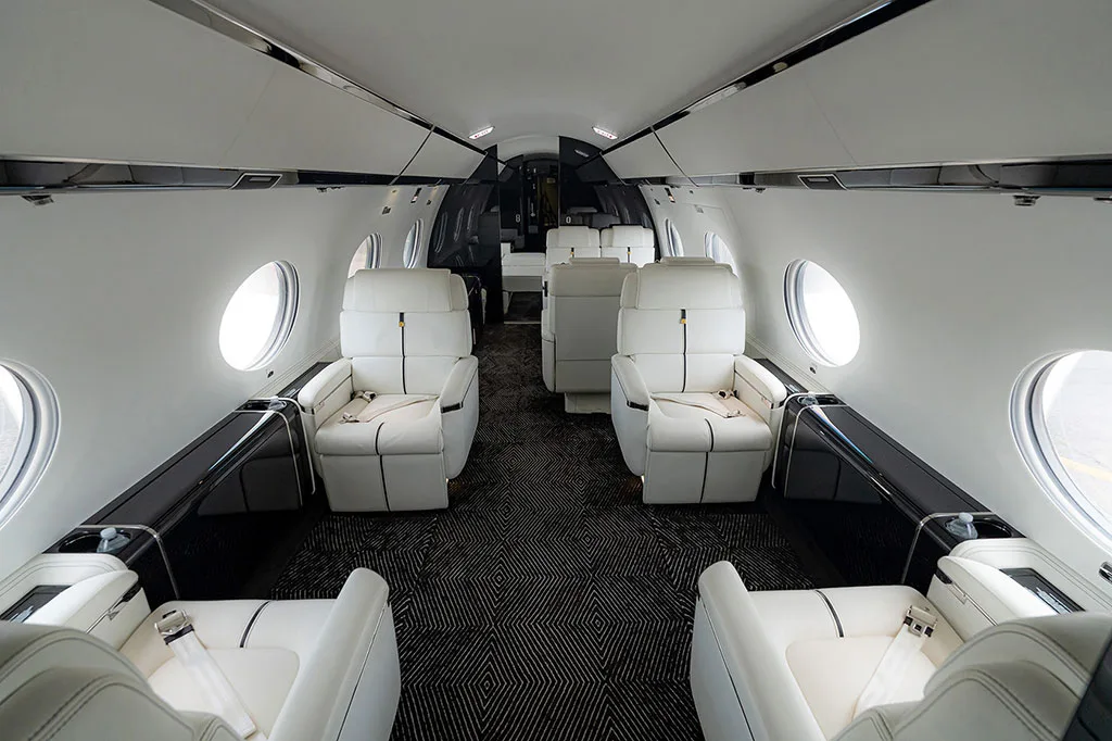 The luxurious cabin interior of a business jet featuring white leather seats and dark patterned carpet.