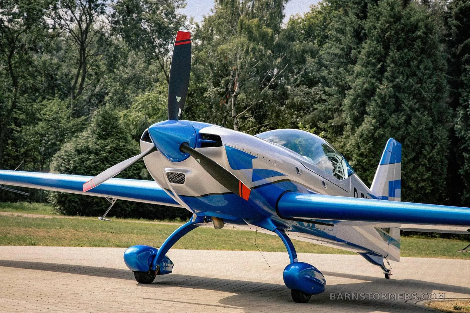 A silver and blue Extra 330SC aircraft is parked on a paved surface with trees in the background.