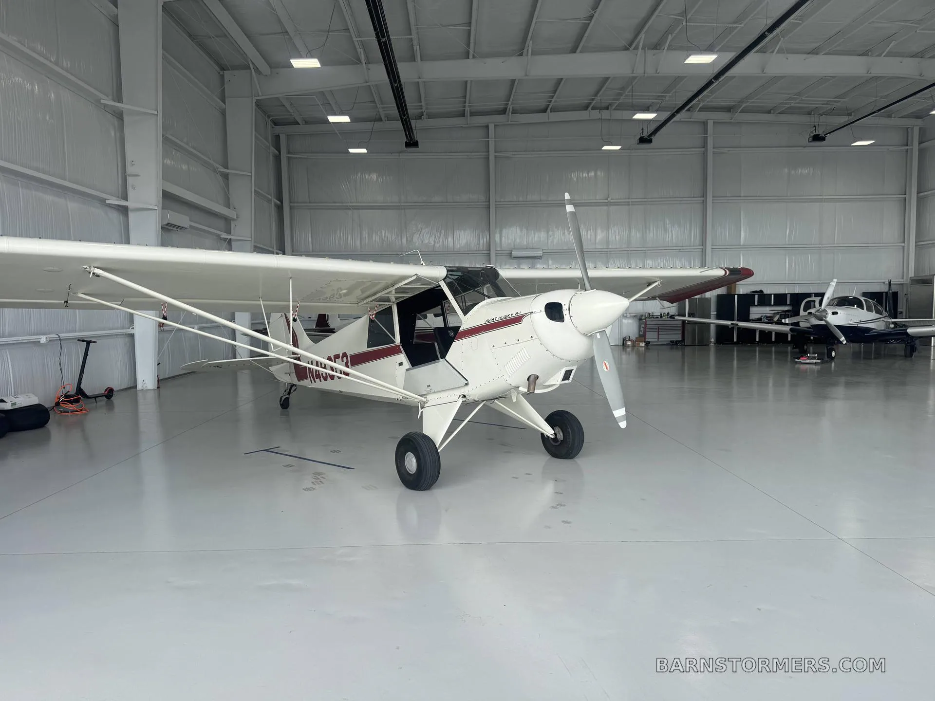 A white and red Aviat Husky A-1 aircraft is parked in a brightly lit hangar, viewed from the front-right.