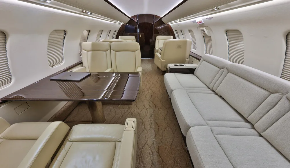 A view inside the private jet cabin showing a dark wood table, a light grey sofa, and beige individual seats.