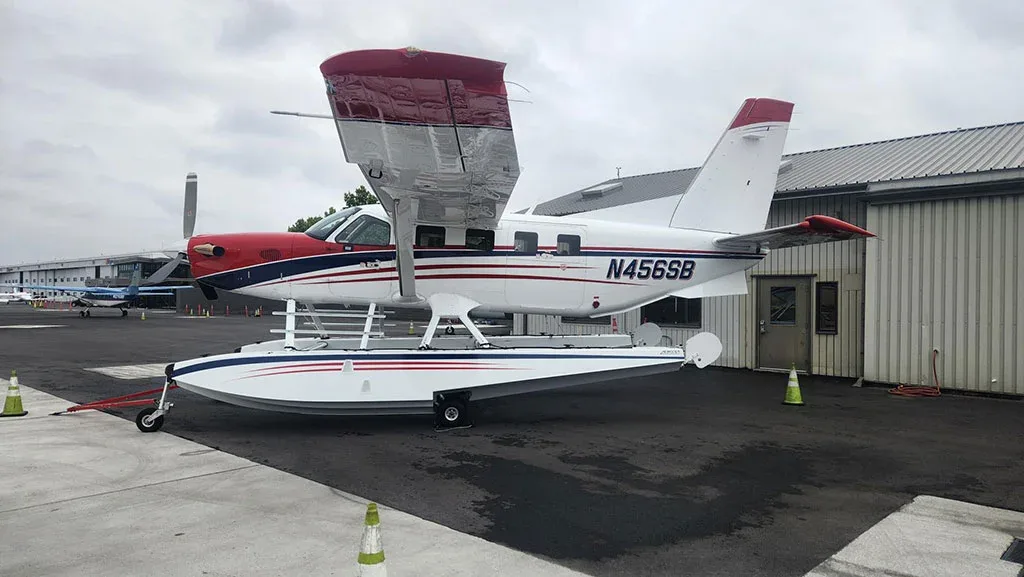 A three-quarter rear view of a red, white, and blue Quest Kodiak 100 on amphibious floats parked on an asphalt apron.