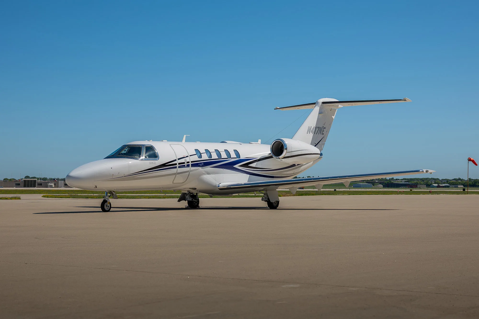 A white Cessna Citation Latitude business jet with blue and grey stripes, tail number N417NE, parked on a tarmac under a clear blue sky.
