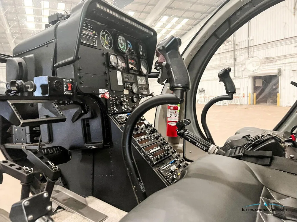 A view of the helicopter cockpit showing the instrument panel, cyclic, collective, and pilot's seat.