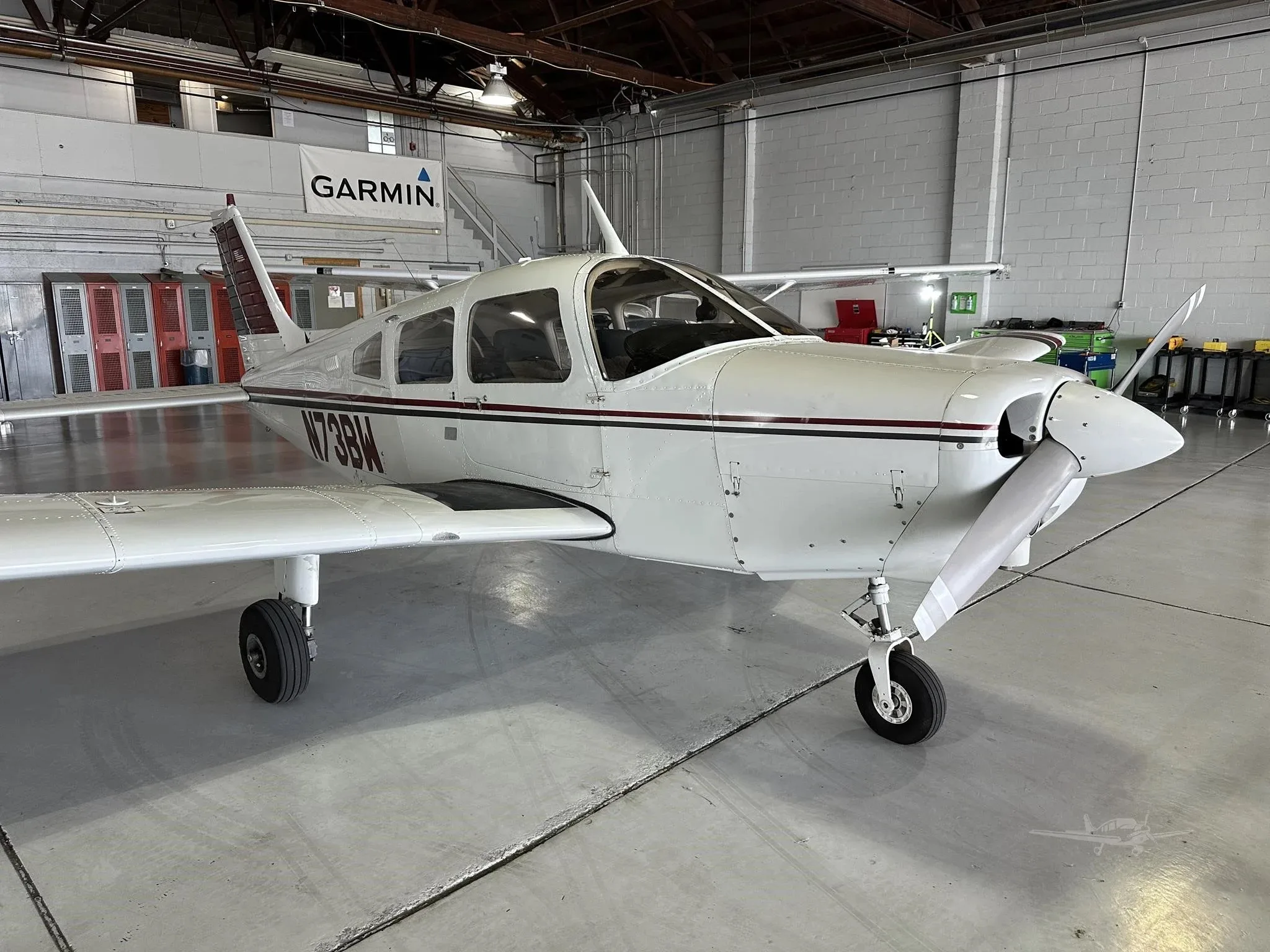 A close-up front-right view of a white single-engine aircraft with maroon and black stripes, registration N73BW, in a hangar.