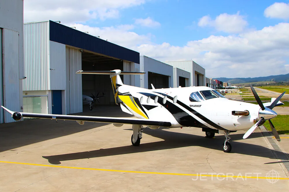 A white, black, and yellow Pilatus PC-12 aircraft parked on an airfield in front of hangars.