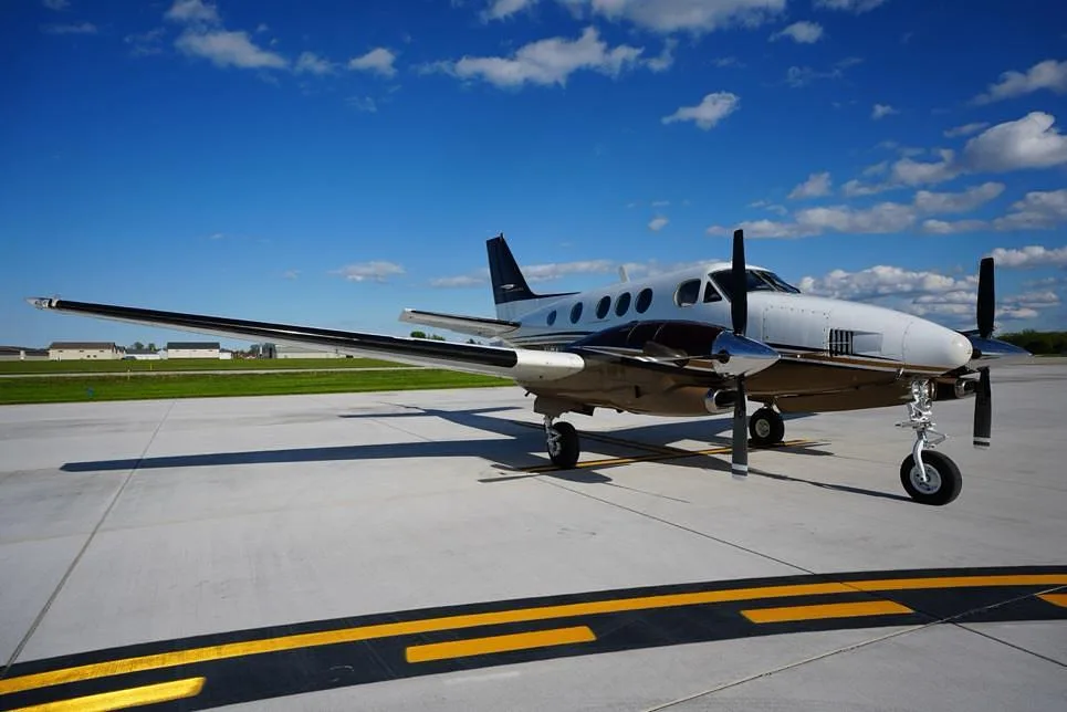 A side view of a white and brown Beechcraft King Air 200 parked on an airfield with yellow taxiway markings.