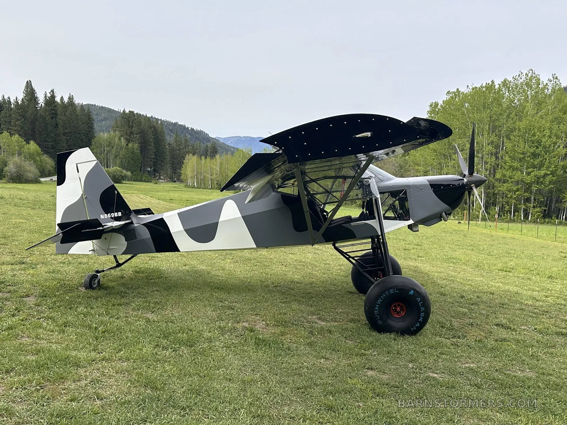 A black, white, and gray camouflage STOL aircraft with large tundra tires is parked on a grassy field with trees and mountains in the background.