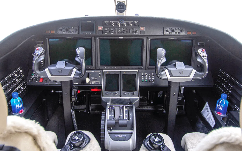 The cockpit of a jet features multiple large display screens, two control yokes, and a central throttle quadrant.