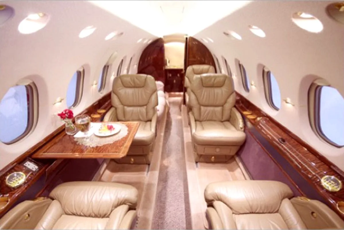 The aircraft cabin features two pairs of opposing tan leather seats with a wooden table set for refreshments in the foreground.