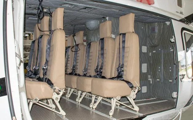 The interior of a helicopter cabin shows a row of tan passenger seats with harnesses.