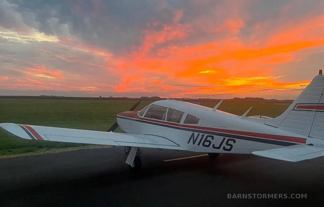A white Piper aircraft with red and black stripes, registration N16JS, parked on a tarmac against a vibrant orange and red sunset sky.